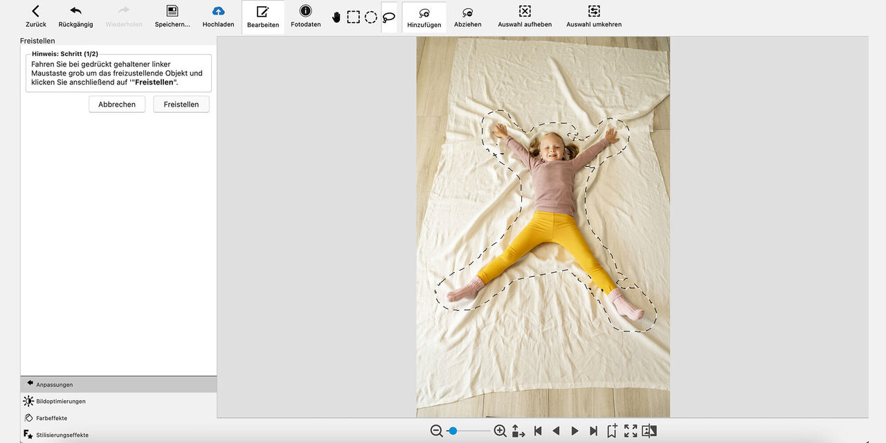 Snímka obrazovky z programu CEWE fotosvet: Dievča leží na podlahe na svetlej rozprestretej deke a usmieva sa do kamery. Dievča má natiahnuté obe ruky a nohy ďaleko od seba. Obrys tela je zhruba načrtnutý prerušovanou čiarou.